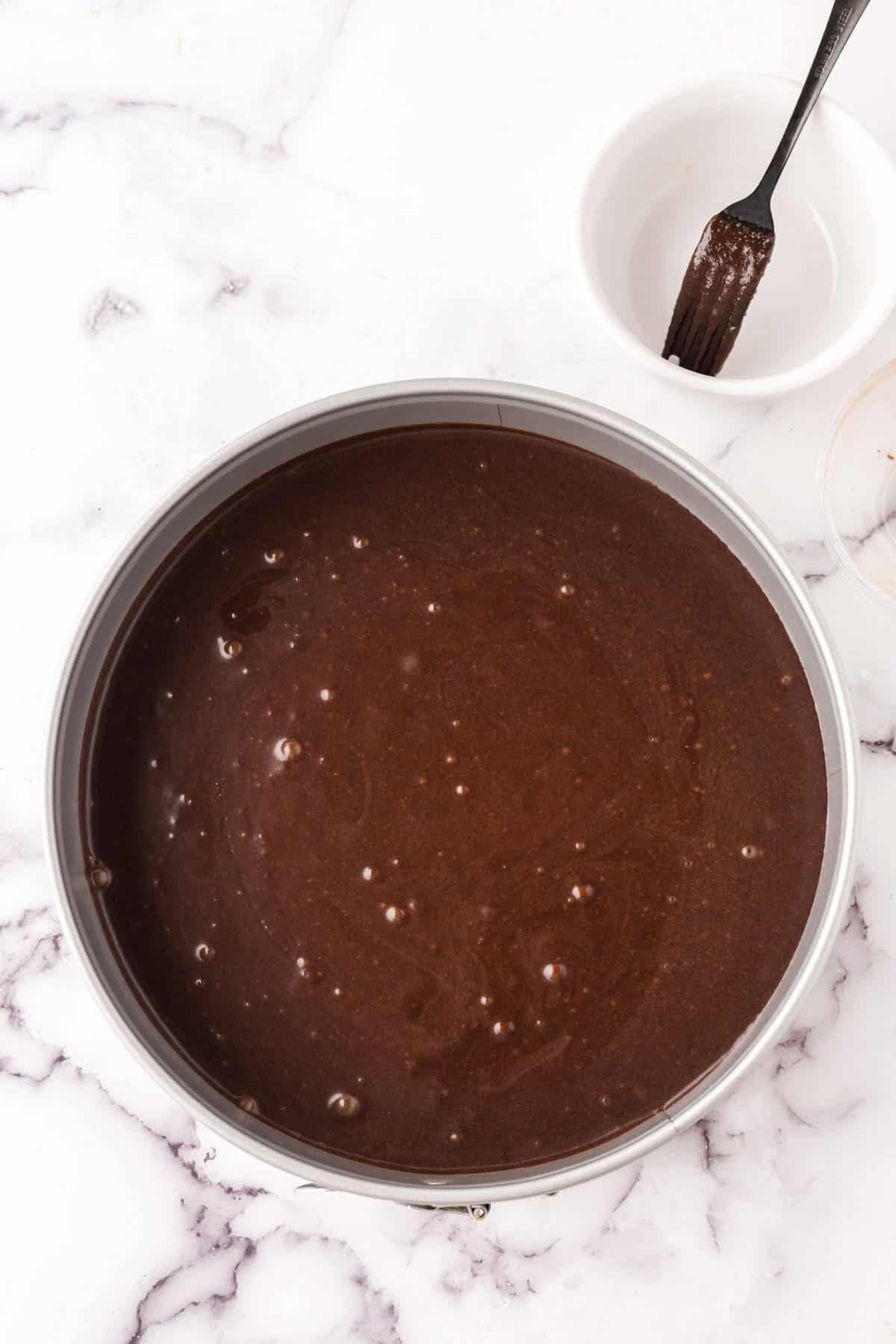 A round cake pan filled with rich flourless chocolate cake batter sits on a white marble surface. Nearby, a white bowl and a fork with chocolate residue hint at the delicious process behind this decadent flourless chocolate cake recipe.