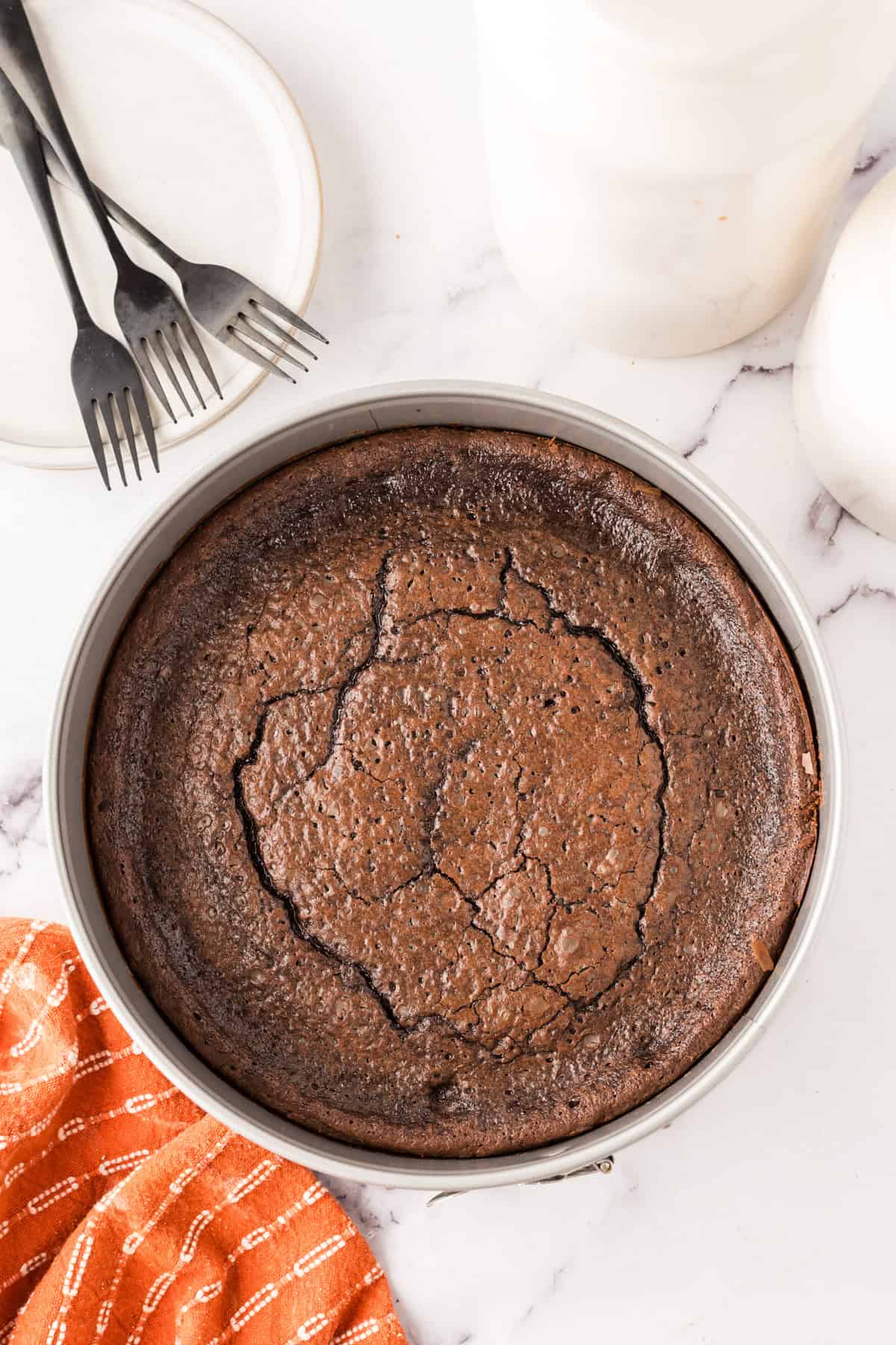 A round flourless chocolate cake with a cracked surface sits in a baking pan on a marble counter. Nearby are an orange textured cloth, two plates with three black forks, and a white bottle. Perfect for trying your next flourless chocolate cake recipe.