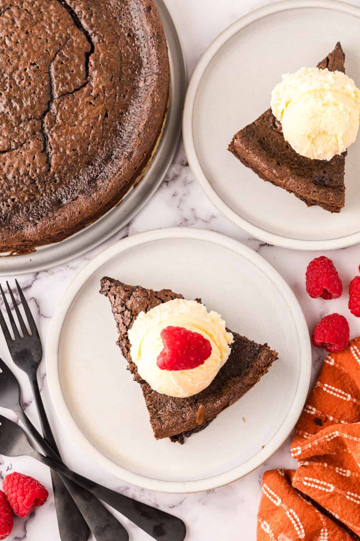 Two plates with slices of flourless chocolate cake, each topped with vanilla ice cream; one scoop is garnished with a raspberry. The whole cake, raspberries, black forks, and an orange cloth are nearby on a marble surface.