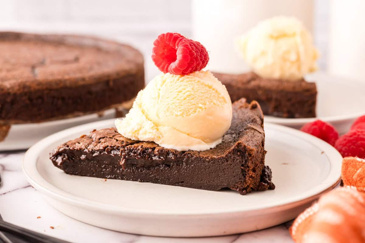 A slice of rich flourless chocolate cake, topped with vanilla ice cream and two raspberries, served on a white plate with more cake and raspberries in the background—an irresistible flourless chocolate cake recipe to try.