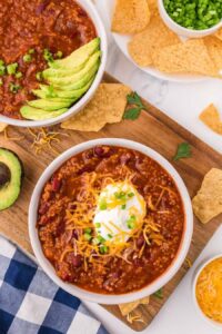 A bowl of chili from a good vegetarian chili recipe is topped with shredded cheese, sour cream, and chopped green onions. It sits on a wooden board next to tortilla chips, avocado slices, and another bowl of chili.