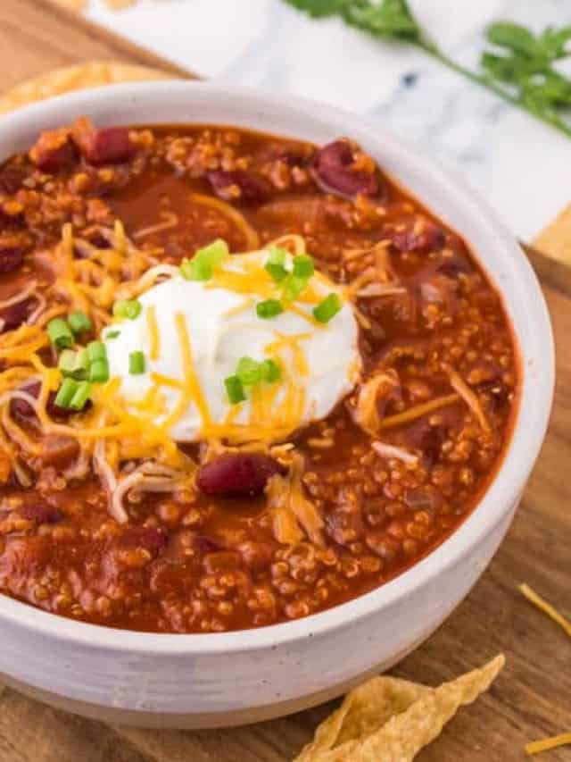 A bowl of good vegetarian chili recipe topped with shredded cheese, a dollop of sour cream, and chopped green onions, served on a wooden board with tortilla chips on the side.