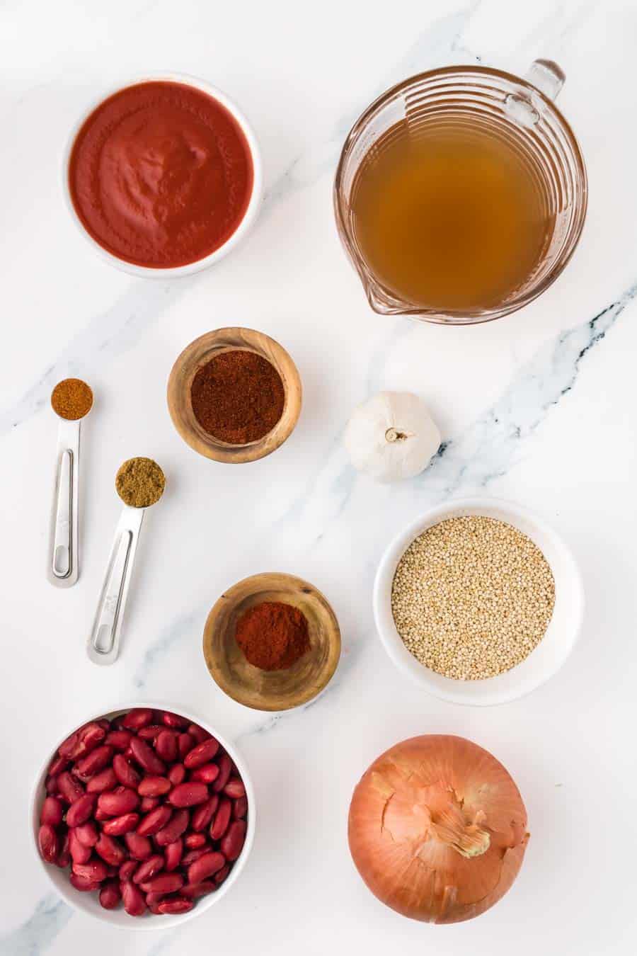 Assorted chili ingredients on a white surface, perfect for a good vegetarian chili recipe—featuring tomato sauce, vegetable broth, garlic, dry quinoa, yellow onion, kidney beans, and small bowls and spoons filled with spices.