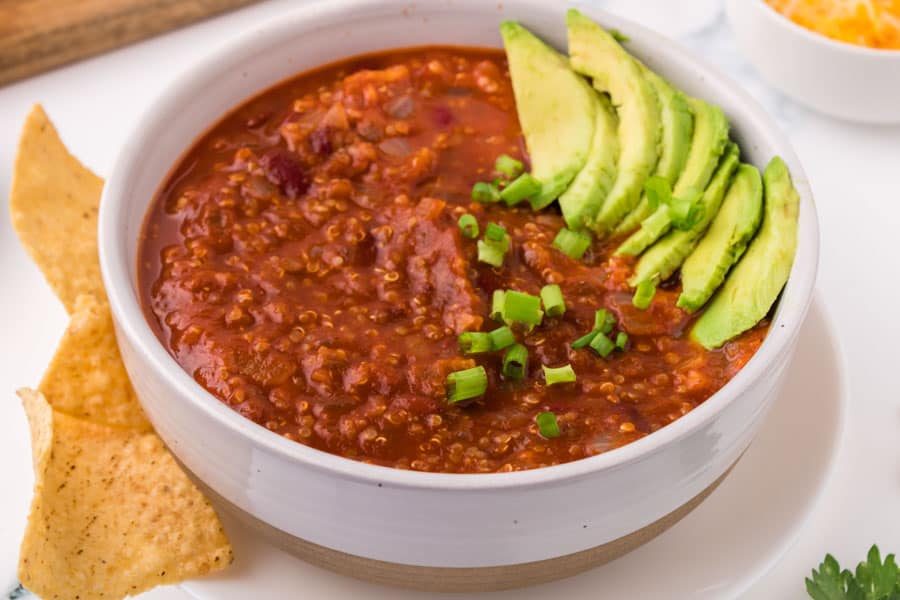 A bowl of good vegetarian chili recipe topped with sliced avocado and chopped green onions, served with tortilla chips on the side.