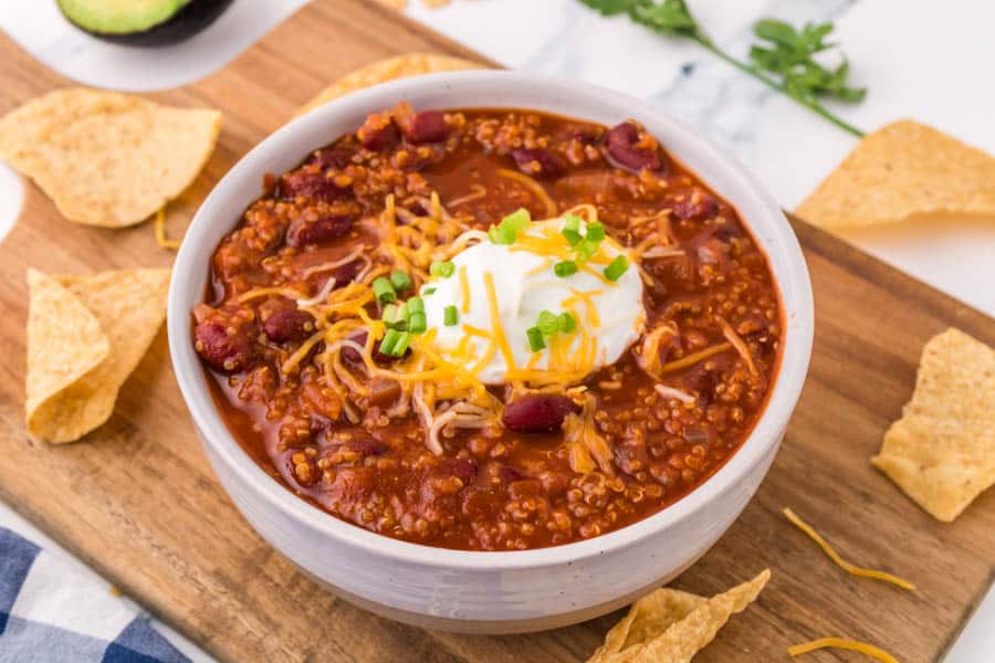 A bowl of good vegetarian chili recipe topped with shredded cheese, sour cream, and chopped green onions sits on a wooden board, surrounded by tortilla chips.