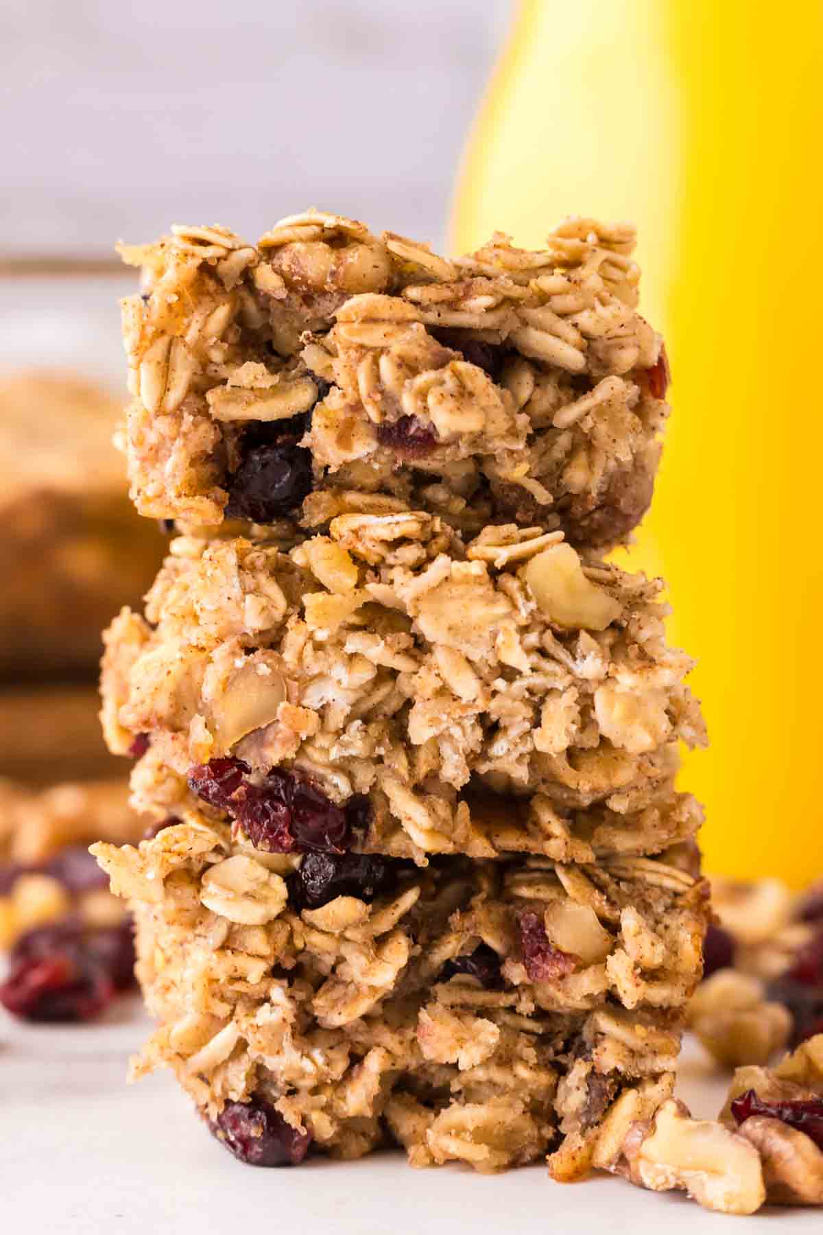 A stack of three homemade oat breakfast bars with visible oats, nuts, and dried cranberries, set against a blurred yellow background.
