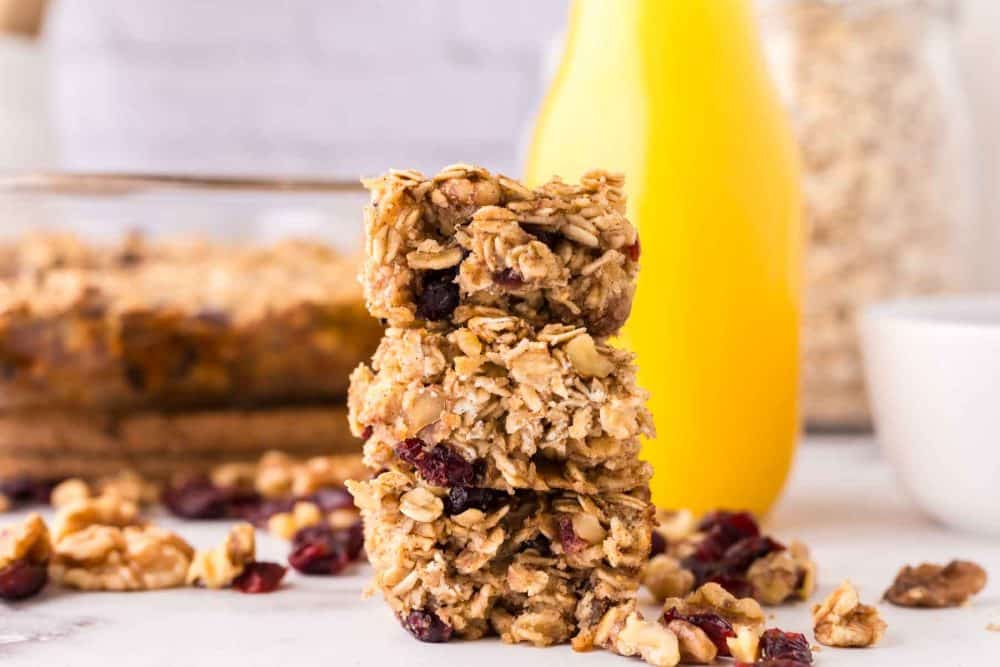 Three stacked oat breakfast bars with dried cranberries and walnuts on a white surface, surrounded by scattered nuts and fruit, with a yellow bottle and a glass dish in the background.