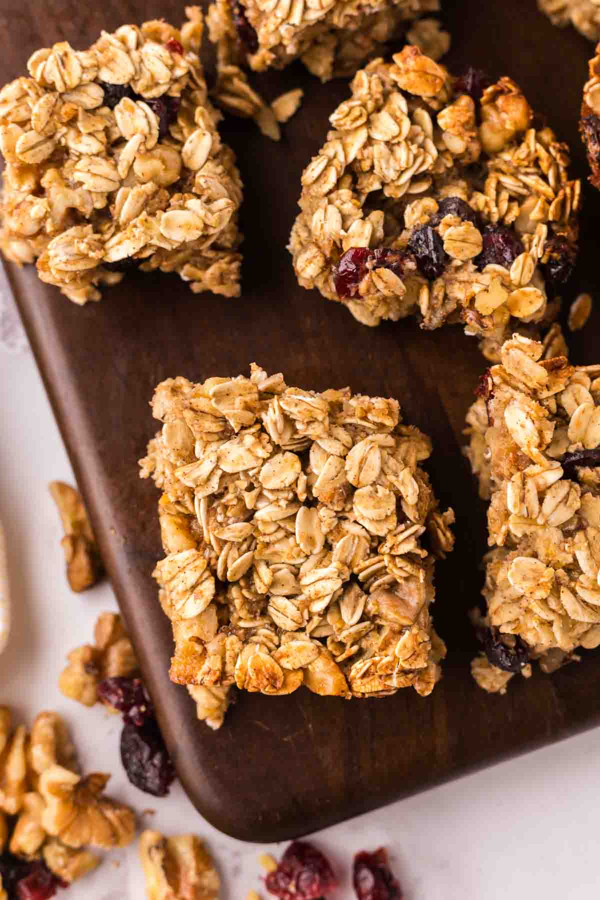 Several oat breakfast bars with dried fruit and nuts are arranged on a dark wooden board, with a few crumbs and loose ingredients scattered nearby.