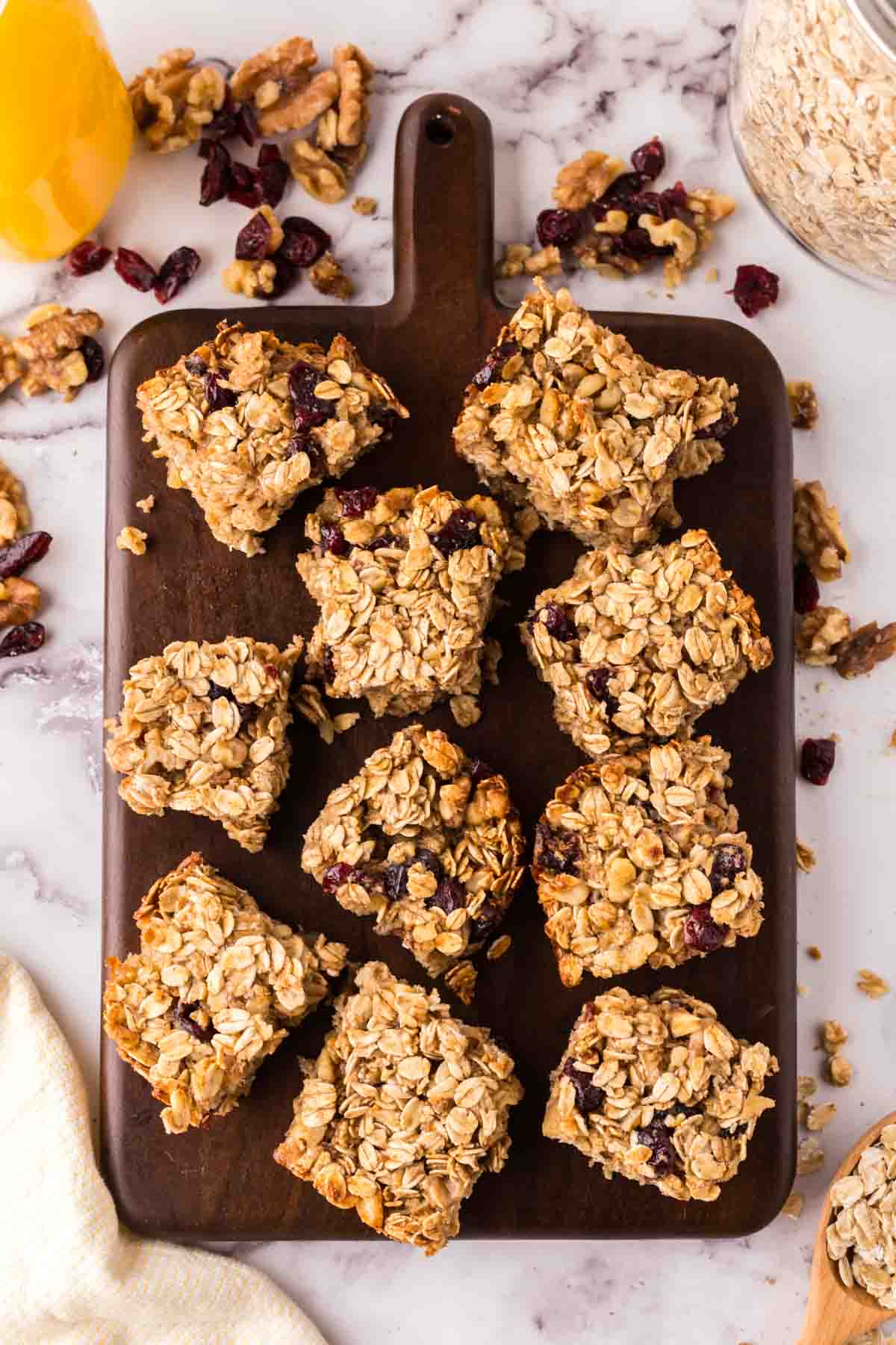 A dark wooden board holds several square oat breakfast bars with visible dried cranberries. The board rests on a marble surface scattered with oats, nuts, and a nearby jar of oats.