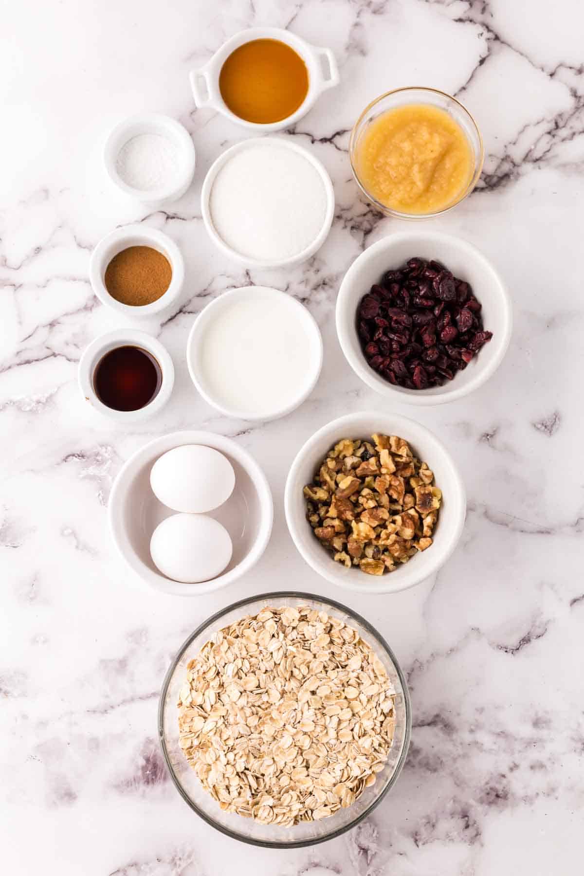 Various baking ingredients arranged on a marble surface for oat breakfast bars, including rolled oats, eggs, walnuts, dried cranberries, milk, applesauce, sugar, vanilla extract, spices, baking powder, and oil.