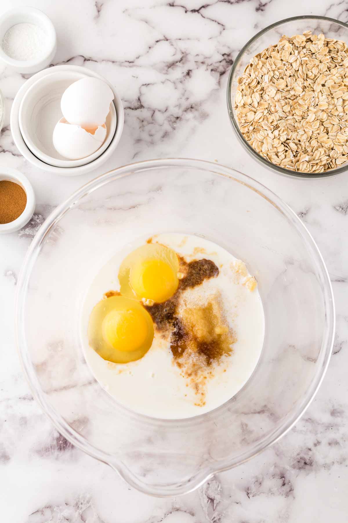 A clear mixing bowl with eggs, milk, brown sugar, and vanilla sits beside oats for oat breakfast bars, two eggs in a smaller bowl, and small bowls of spices and salt on a marble countertop.