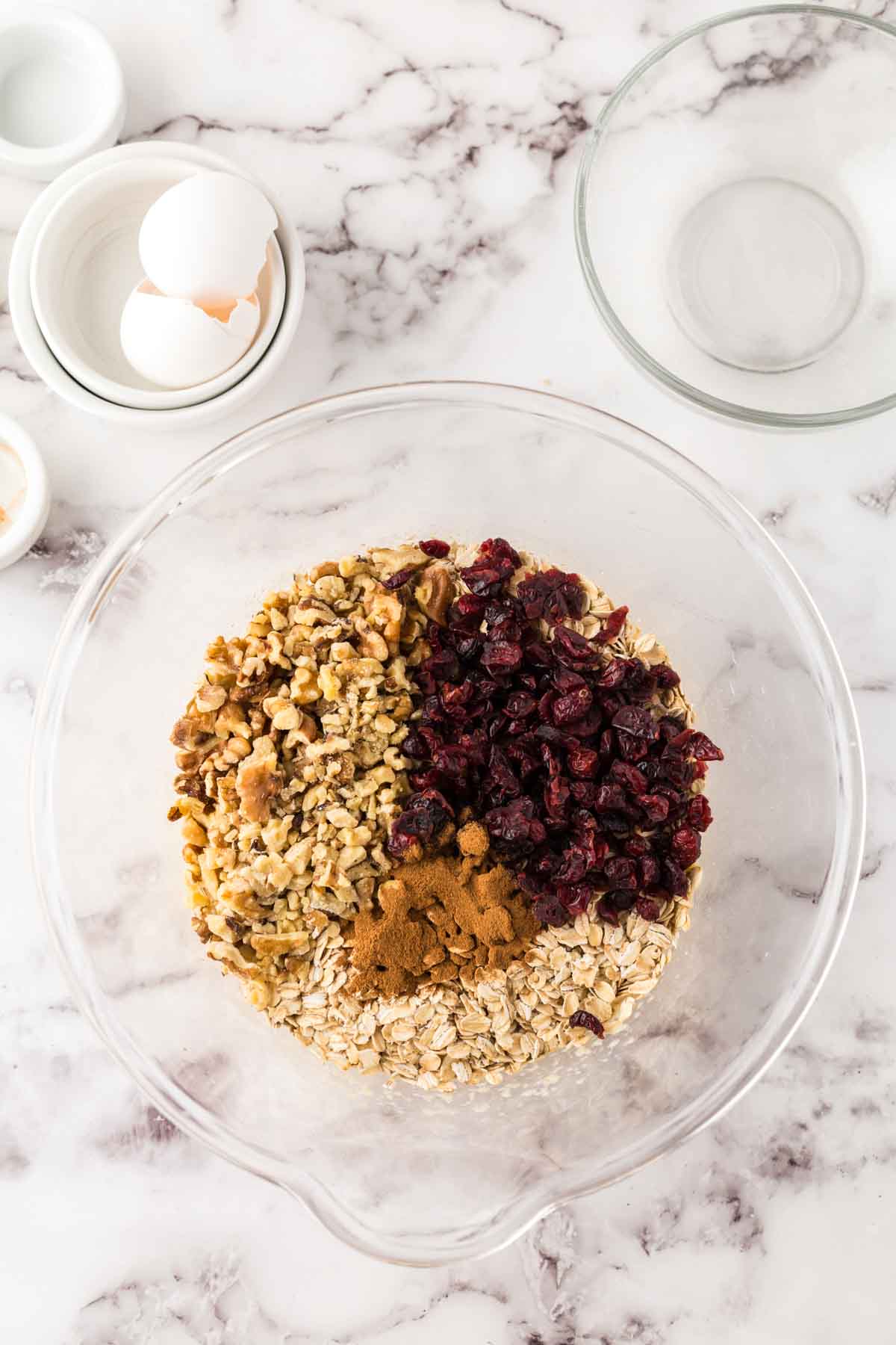 A glass mixing bowl with oats, chopped walnuts, dried cranberries, and cinnamon sits on a marble countertop—perfect ingredients for homemade oat breakfast bars—surrounded by empty bowls and eggshells.