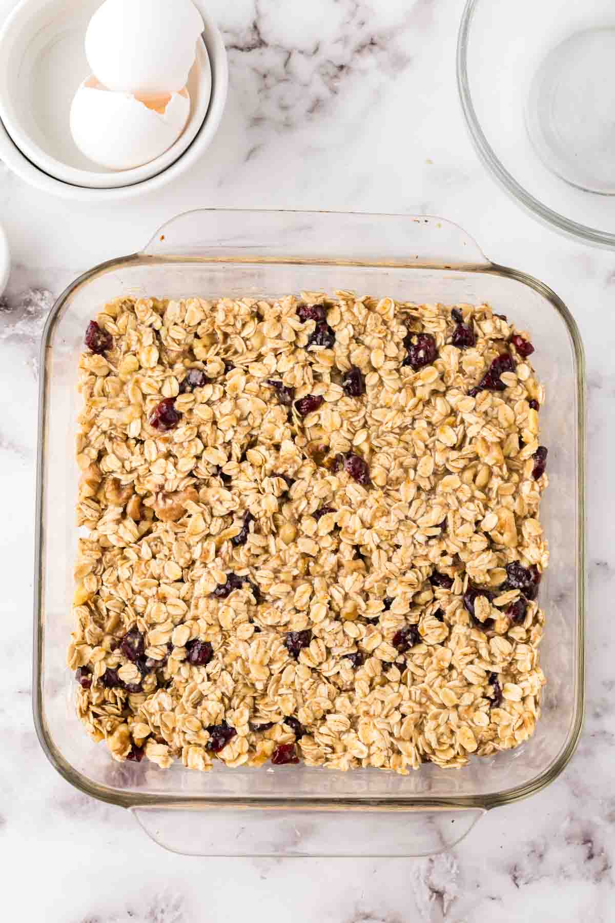 A square glass baking dish filled with baked oatmeal, perfect for cutting into oat breakfast bars, sits on a marble countertop. Scattered dried berries stud the oats, while cracked eggshells and an empty glass bowl rest nearby.