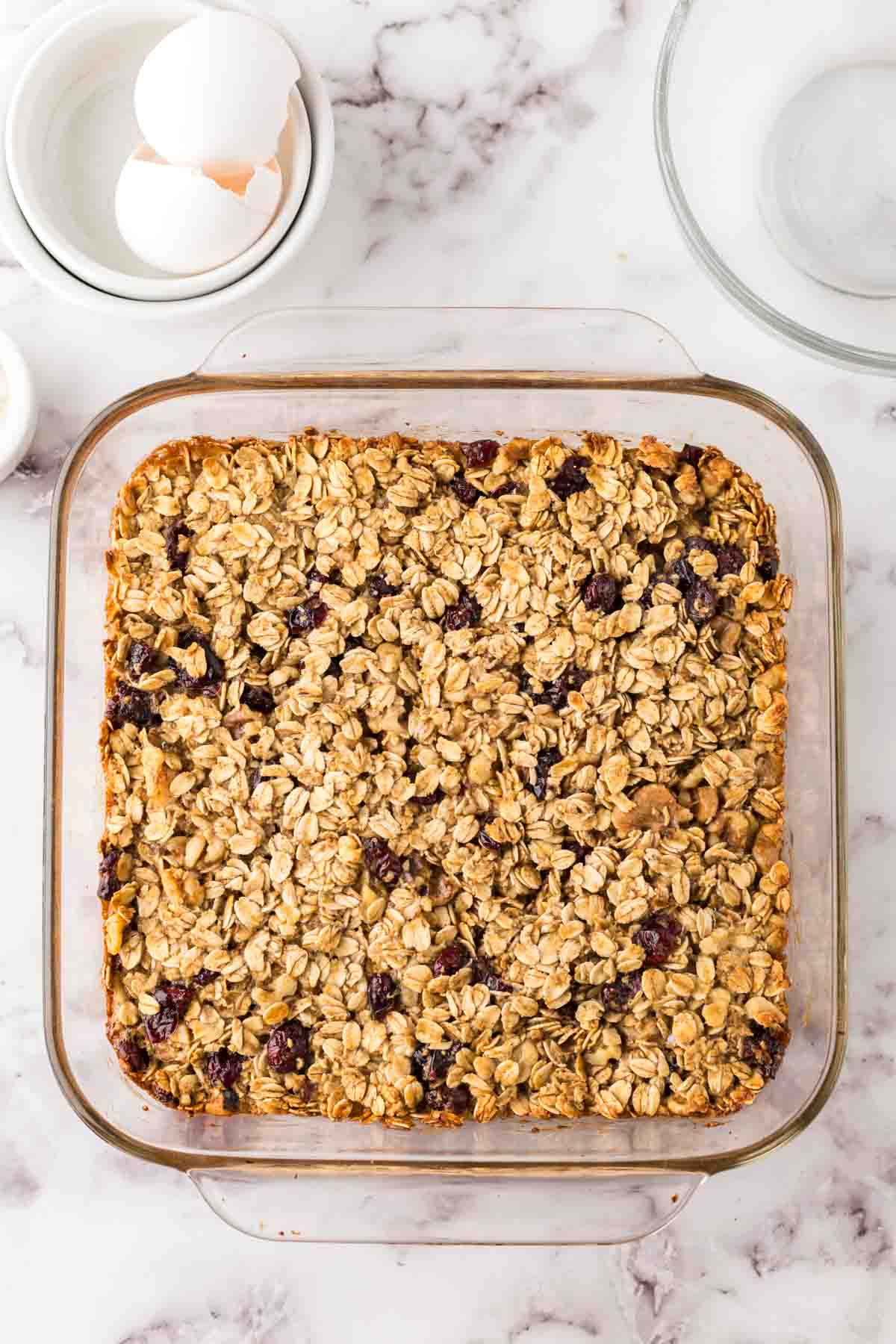 A glass baking dish filled with oat breakfast bars topped with oats and berries sits on a marble countertop next to a bowl of eggs and a clear empty bowl.