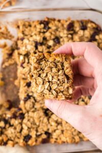 A hand holds a square oat breakfast bar with visible oats and dried fruit, above a glass baking dish filled with more oat breakfast bars.