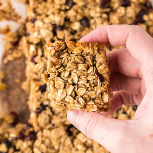 A hand holds a square oat breakfast bar with visible oats and dried fruit, above a glass baking dish filled with more oat breakfast bars.