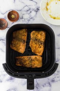 Three seasoned salmon fillets in air fryer basket await cooking. Small bowls of spices and a plate with marinade residue are set beside the fryer on a marble countertop.
