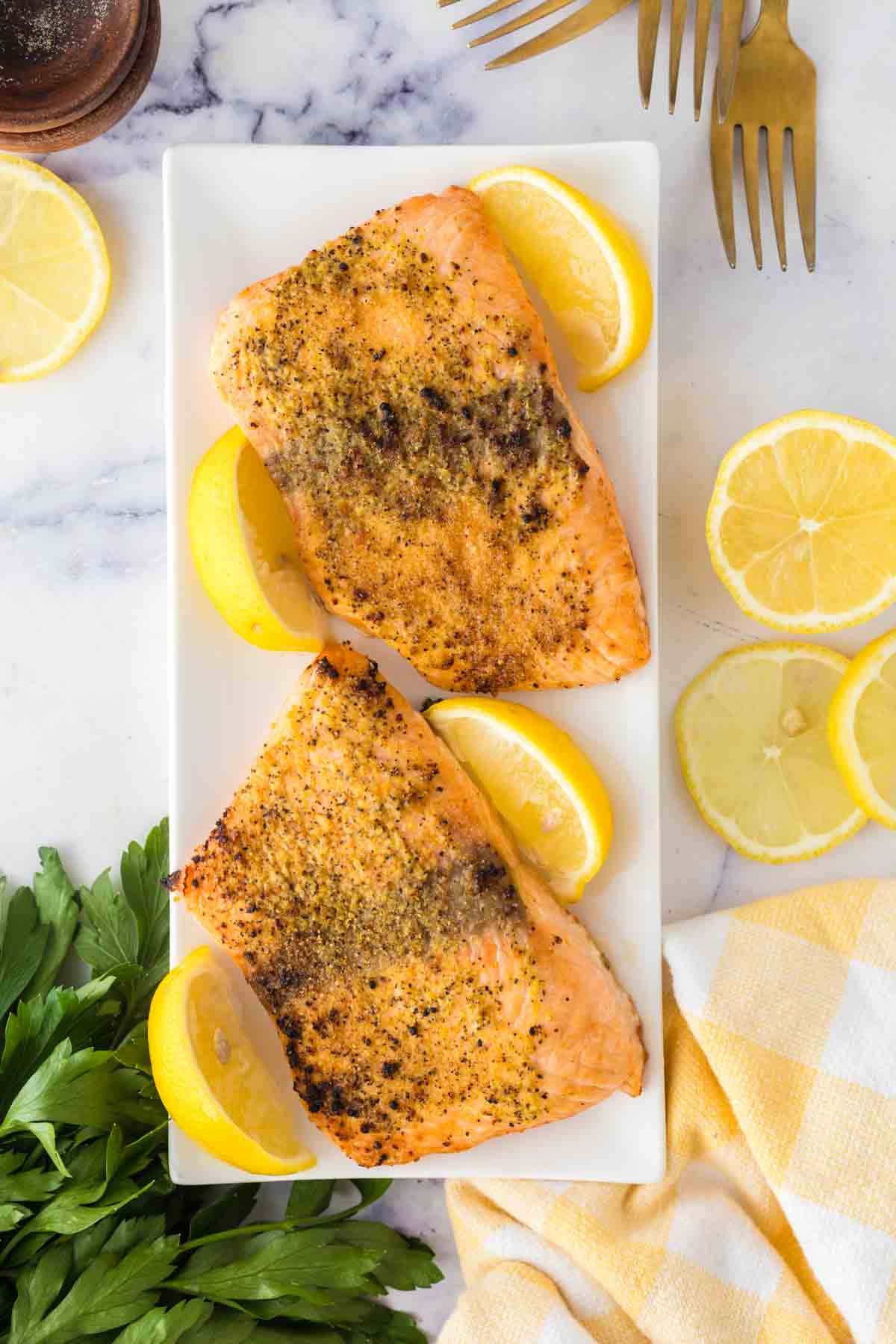 Two seasoned salmon fillets in air fryer, baked to perfection, rest on a rectangular white plate with lemon wedges. Slices of lemon, fresh parsley, gold forks, and a yellow-checked napkin surround the plate on a marble surface.