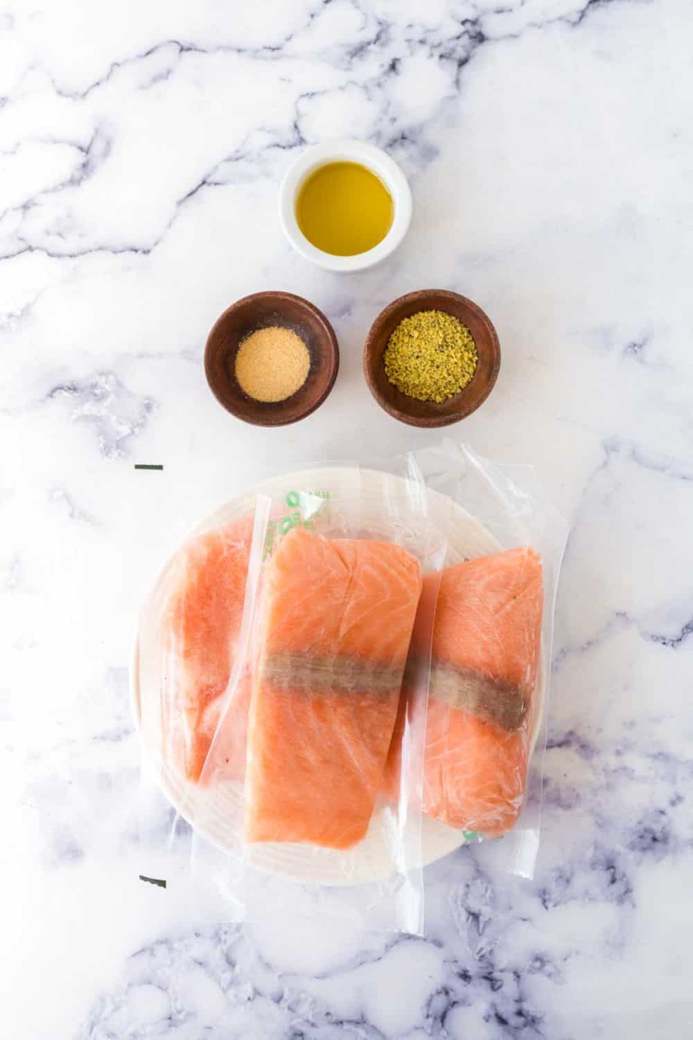 Two vacuum-sealed salmon fillets on a plate, ready to become salmon fillets in air fryer, with small bowls of olive oil, seasoning, and garlic powder above—all set on a white marble surface.