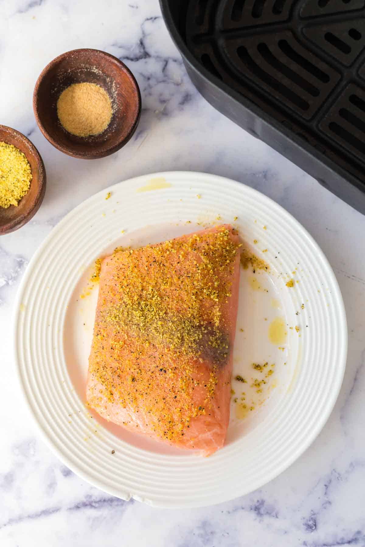 A raw salmon fillet seasoned with spices sits on a white plate beside two small bowls of seasoning and an empty air fryer basket, perfect for making salmon fillets in air fryer, all arranged on a marble surface.