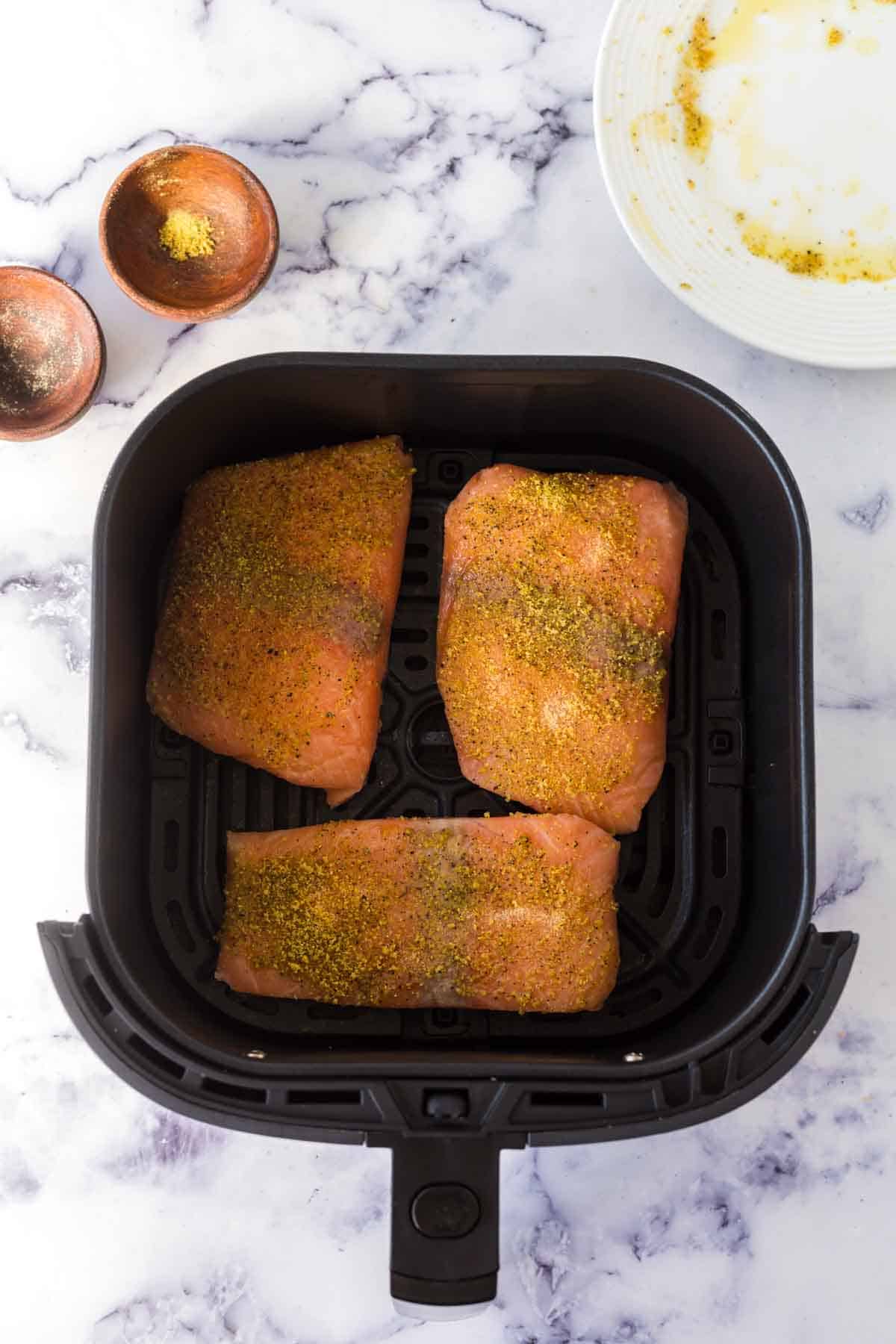 Three raw salmon fillets in air fryer basket are seasoned with spices and ready for cooking. Small bowls of seasoning and a white plate with spice residue sit nearby on a marble countertop.