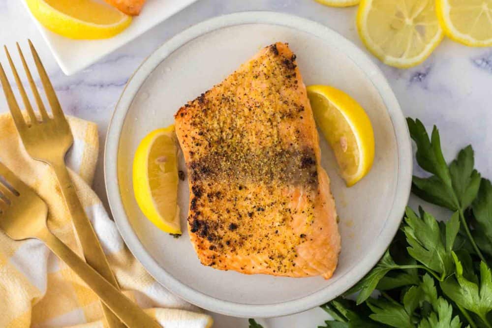 A cooked salmon fillet seasoned with spices sits on a plate with two lemon wedges, surrounded by fresh parsley, lemon slices, two gold forks, and a yellow-striped napkin—perfect presentation for salmon fillets in air fryer.
