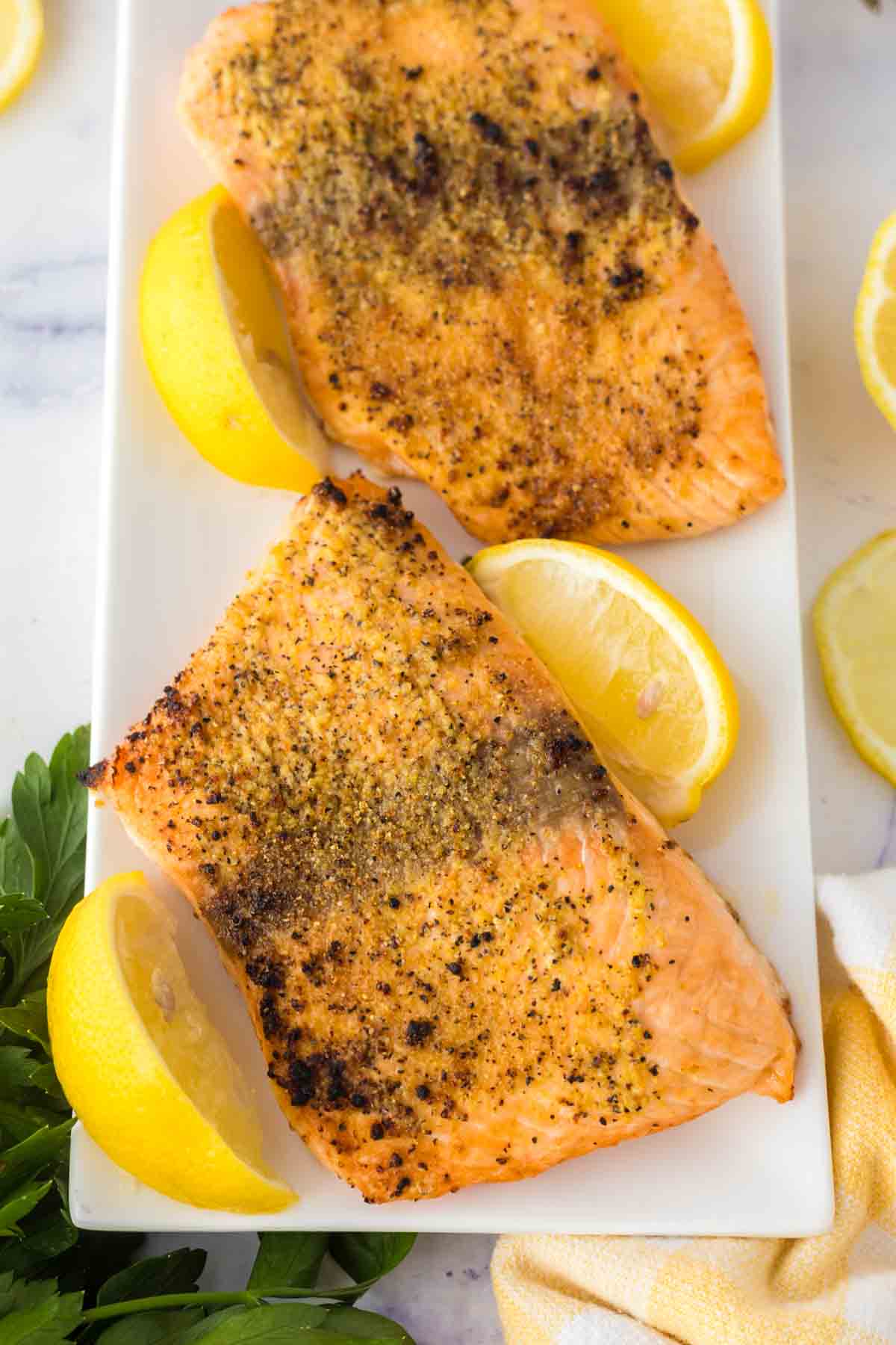Two seasoned salmon fillets in air fryer, baked to perfection and served on a white rectangular plate with lemon wedges, garnished with herbs. The salmon has a golden, slightly crispy top.