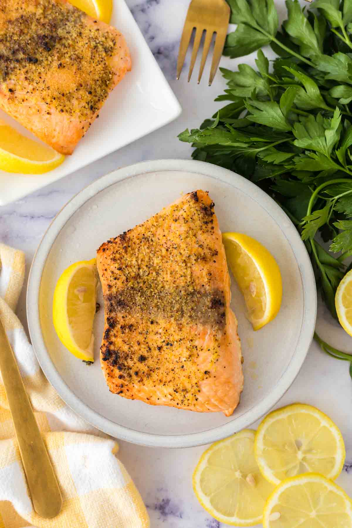 A plate with a seasoned, baked salmon fillet garnished with two lemon wedges. Fresh parsley, lemon slices, a gold fork, and another plate with salmon fillets in air fryer style are nearby on a marble surface.