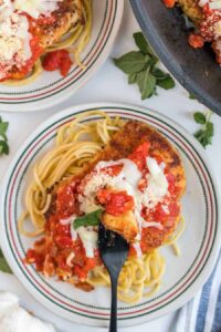 A plate of skillet chicken parmesan features breaded chicken topped with tomato sauce and melted cheese over spaghetti. A fork holds a piece of the chicken, garnished with fresh herbs. Another plate and a pan are partially visible nearby.