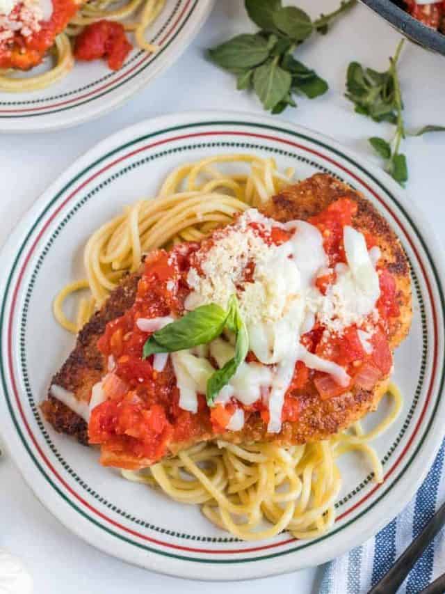 A plate of skillet chicken parmesan sits atop spaghetti, smothered in marinara sauce, melted cheese, grated Parmesan, and a basil garnish. Fresh herbs and another plate are visible in the background.