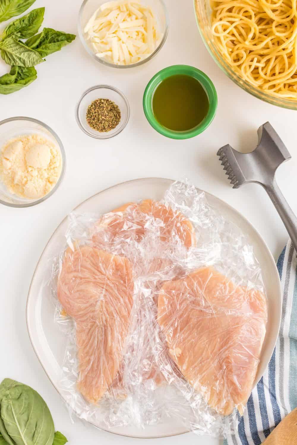 Raw chicken breasts covered in plastic wrap sit on a plate next to a meat tenderizer, spices, cheese, olive oil, fresh basil, and a bowl of cooked spaghetti—perfect for preparing homemade skillet chicken parmesan.