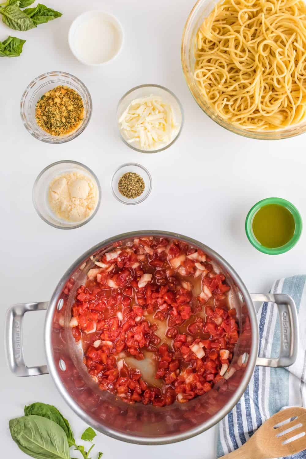 A pot of chopped tomatoes and onions on a white surface is surrounded by bowls of cooked spaghetti, shredded cheese, seasonings, grated cheese, olive oil, and fresh basil leaves—perfect ingredients for skillet chicken parmesan. A blue-striped towel and wooden spoon are beside them.