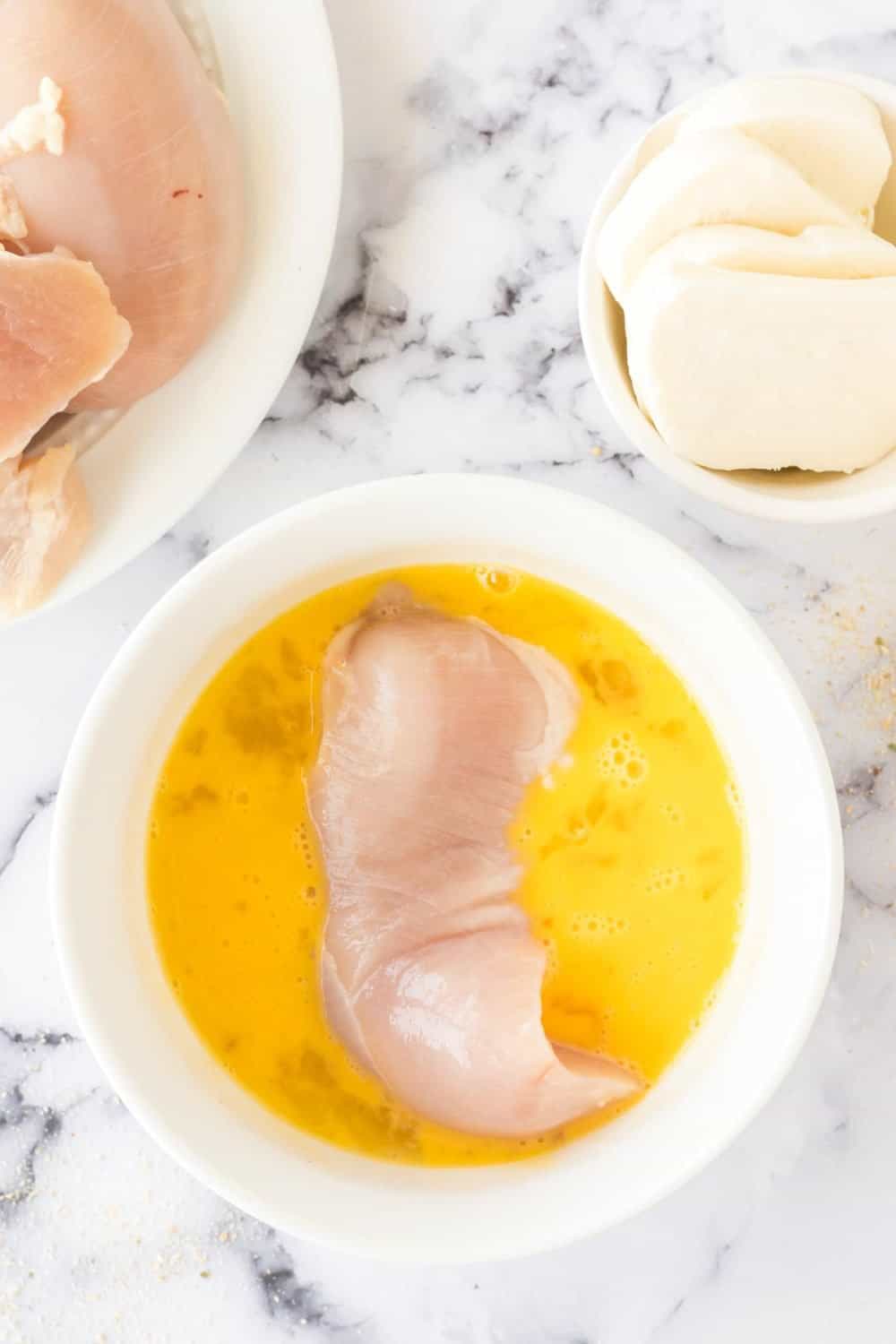 A raw chicken breast sits in a bowl of beaten eggs on a marble countertop, ready for skillet chicken parmesan. Nearby, a plate holds more raw chicken and a small bowl contains slices of mozzarella cheese.