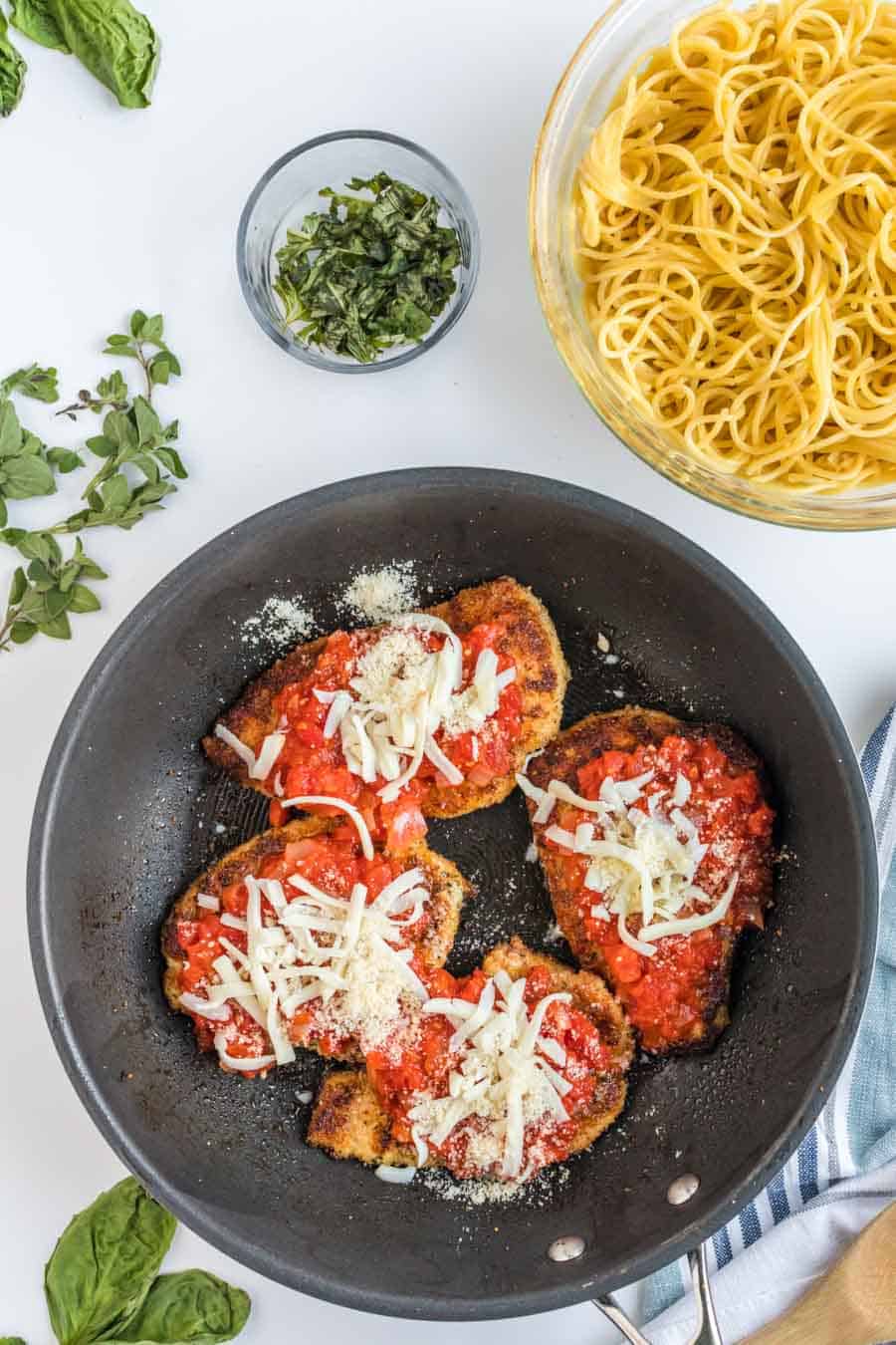 A skillet chicken parmesan with breaded chicken breasts topped with tomato sauce and shredded cheese sits next to a bowl of cooked spaghetti, fresh herbs, and a small dish of chopped basil on a white surface.