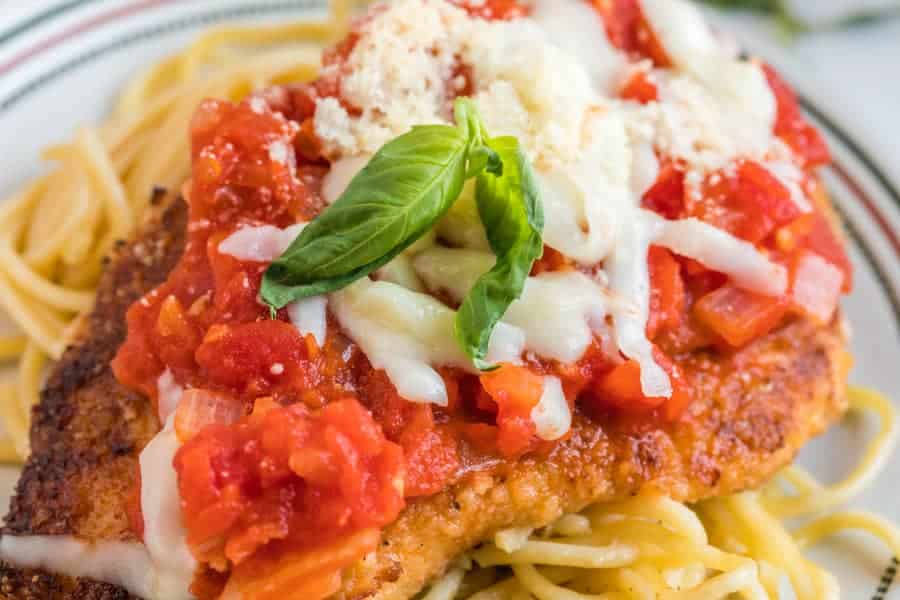 A close-up of skillet chicken parmesan: a breaded chicken cutlet topped with marinara sauce, melted cheese, grated parmesan, and a fresh basil leaf, served on a bed of spaghetti pasta.