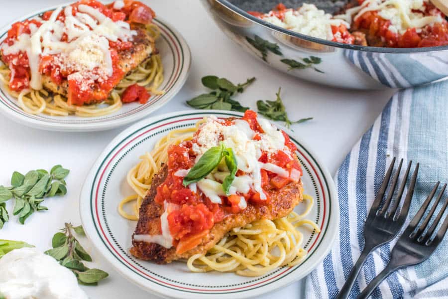 A plate of skillet chicken parmesan—breaded chicken over spaghetti with tomato sauce, melted cheese, basil, and parmesan—sits beside fresh herbs, extra food in a bowl, and a blue-striped napkin with two black forks.
