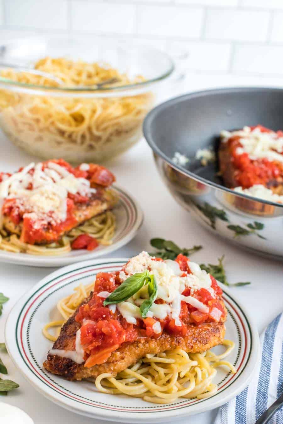 Two plates of skillet chicken parmesan topped with tomato sauce and melted cheese are served over spaghetti. In the background, bowls of plain spaghetti and more skillet chicken parmesan rest on a white countertop.