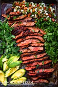Sliced grilled steak, seasoned with Tex Mex seasoning, arranged on a tray with fresh avocado slices, cilantro, and colorful pico de gallo salsa. The vibrant food is neatly displayed from Longbourn Farm.