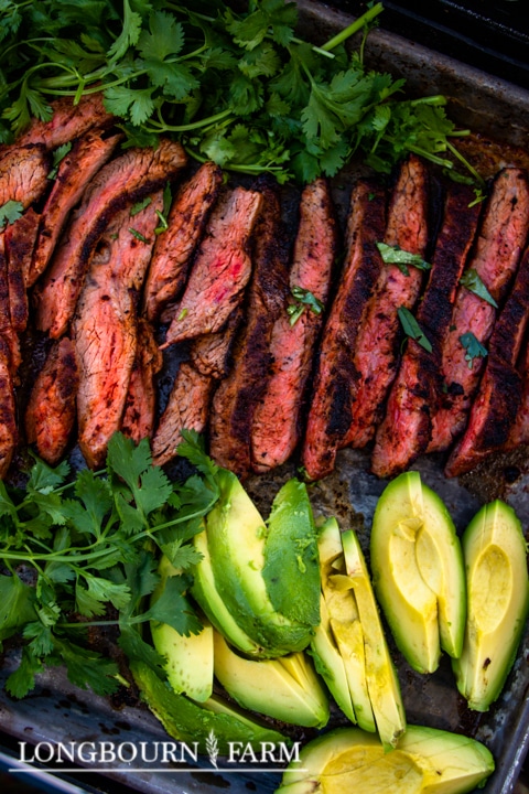 Sliced grilled steak with a charred exterior and pink center, seasoned with Tex Mex seasoning, garnished with fresh cilantro and served alongside sliced avocado on a tray. The image has LONGBOURN FARM text in the corner.