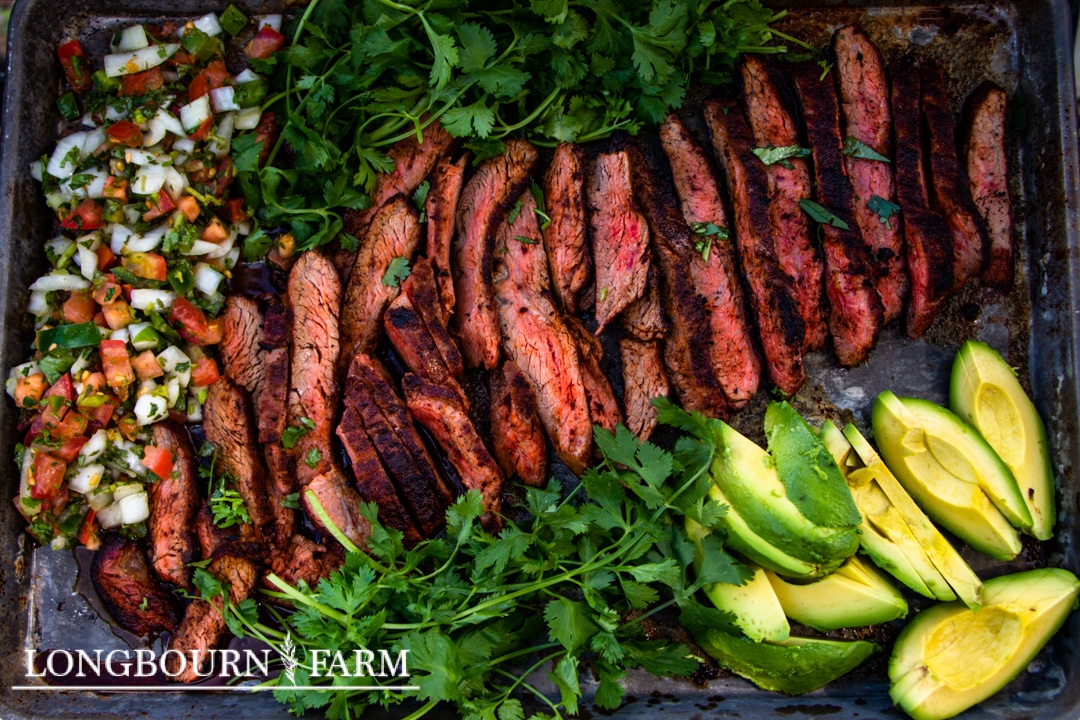 Sliced grilled steak seasoned with Tex Mex seasoning, fresh pico de gallo, avocado slices, and cilantro are arranged on a baking sheet. The medium-rare steak boasts visible grill marks and a sprinkle of herbs.