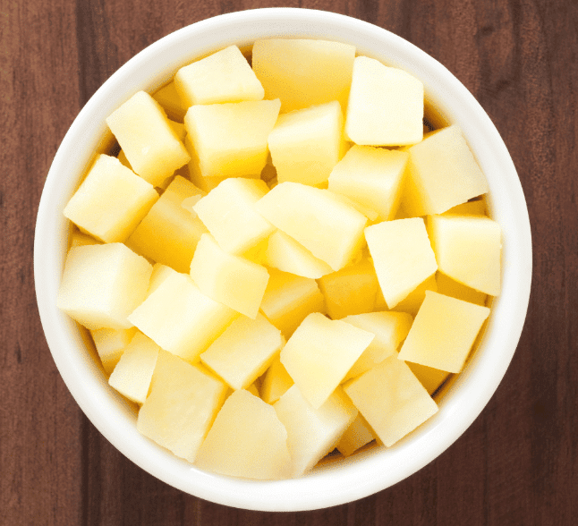 A white bowl filled with evenly cut cubes of raw potato sits on a wooden surface, viewed from above.