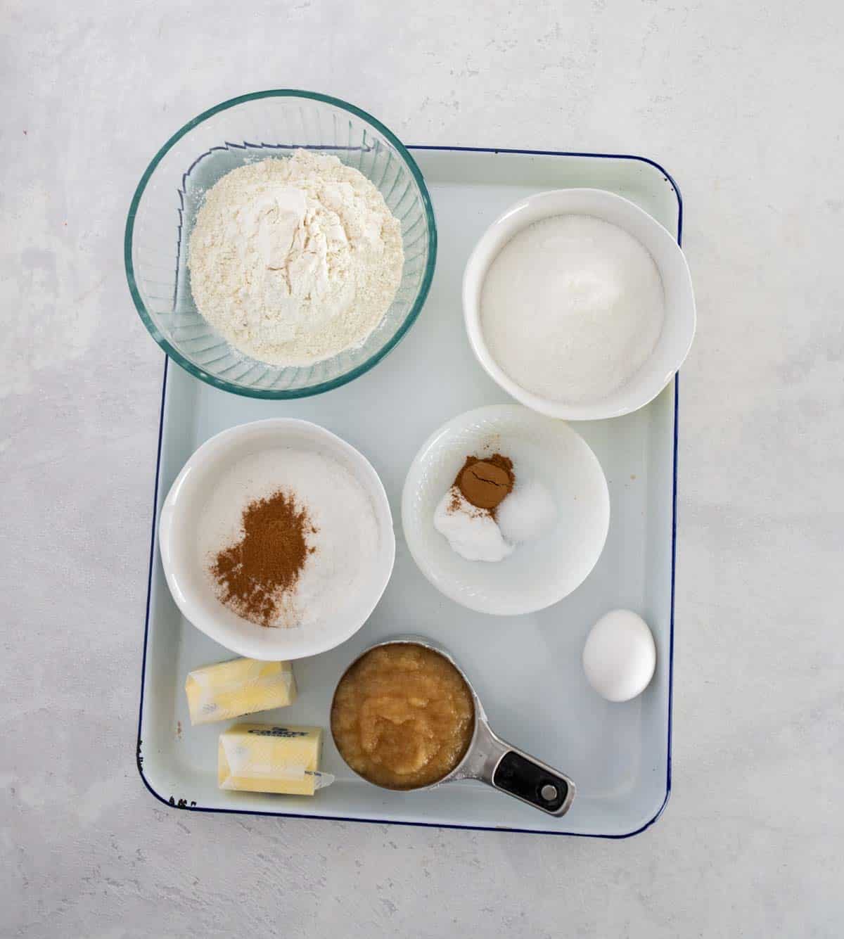 A tray with bowls containing flour, sugar, cinnamon, baking powder, baking soda, salt, butter sticks, applesauce, and a whole egg on a light countertop.