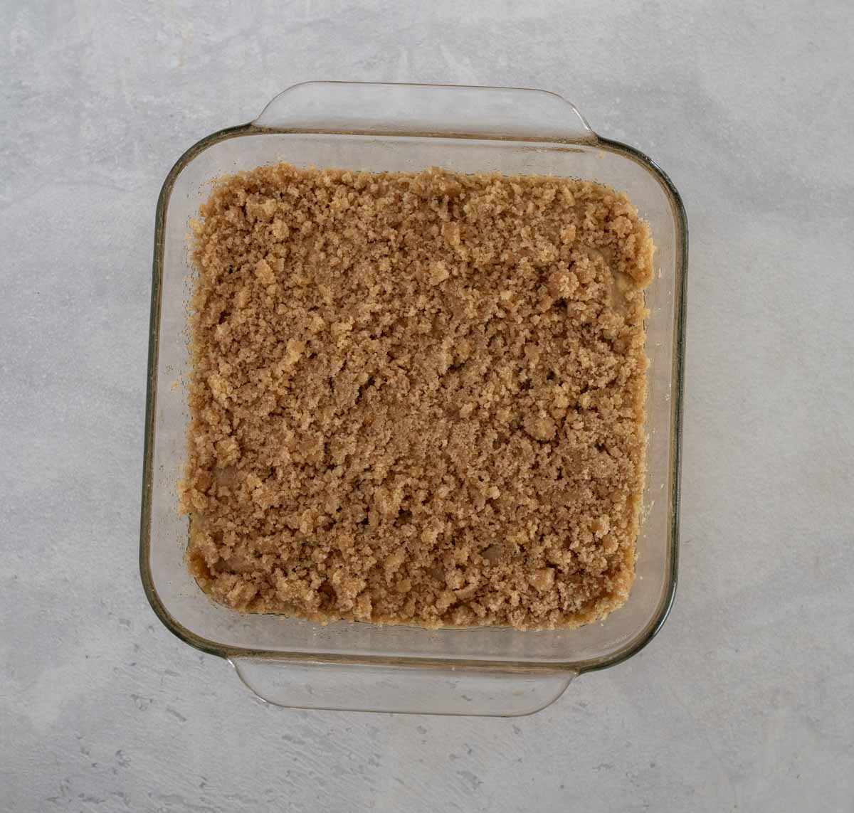 A square glass baking dish filled with a layer of brown, crumbly topping, set on a light gray textured surface.