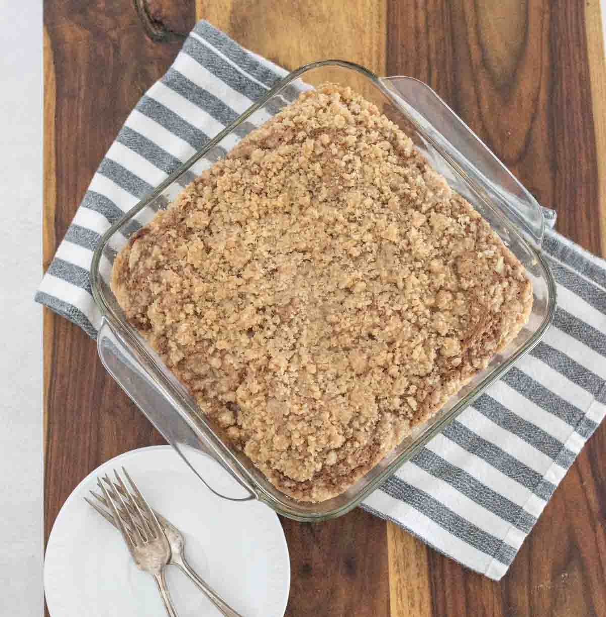 A glass baking dish filled with crumb-topped coffee cake sits on a striped towel on a wooden table. Nearby, two forks rest on a white plate.