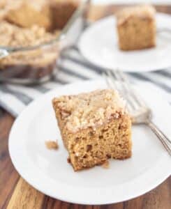 A square piece of crumb-topped coffee cake sits on a white plate with a fork, and another piece is on a plate in the background, next to a glass baking dish on a striped cloth.