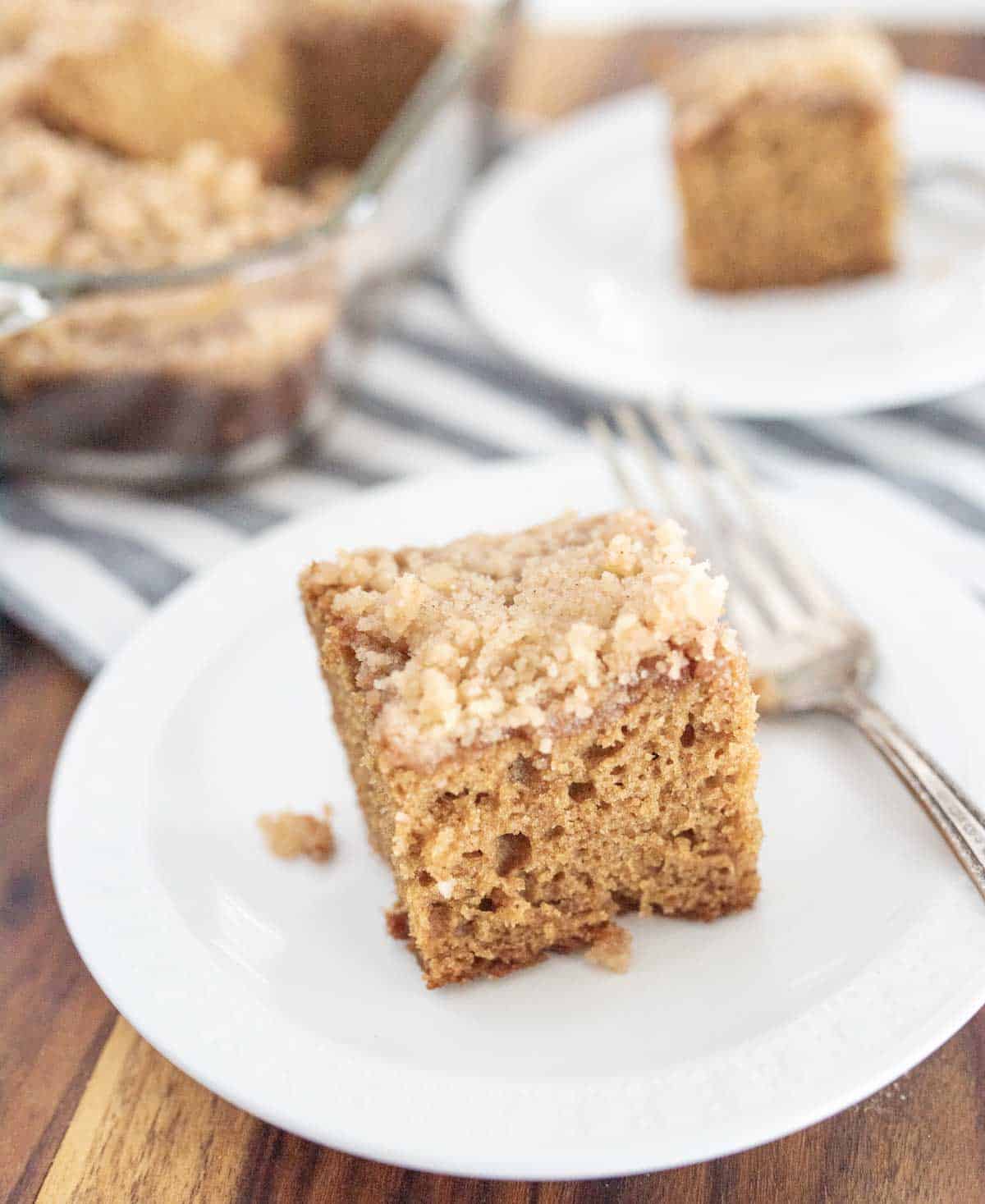 A square piece of crumb-topped coffee cake sits on a white plate with a fork, and another piece is on a plate in the background, next to a glass baking dish on a striped cloth.