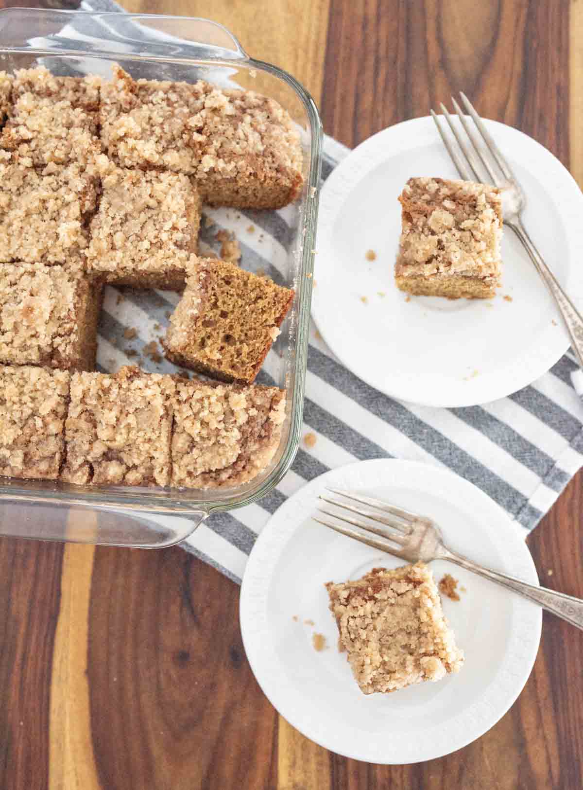 A glass dish of sliced coffee cake with cinnamon crumb topping sits on a striped towel, with two pieces served on white plates, each with a fork, on a wooden table.