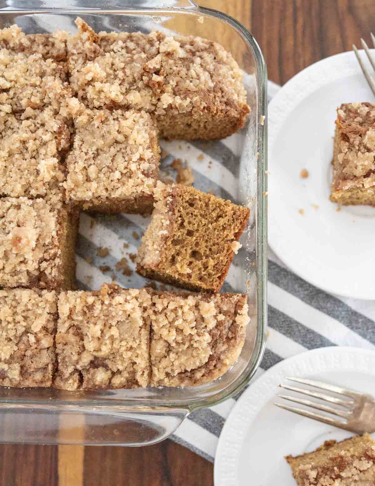 A glass baking dish with sliced pieces of crumb-topped coffee cake, some pieces removed and served on white plates with forks, all on a striped cloth and wooden table.