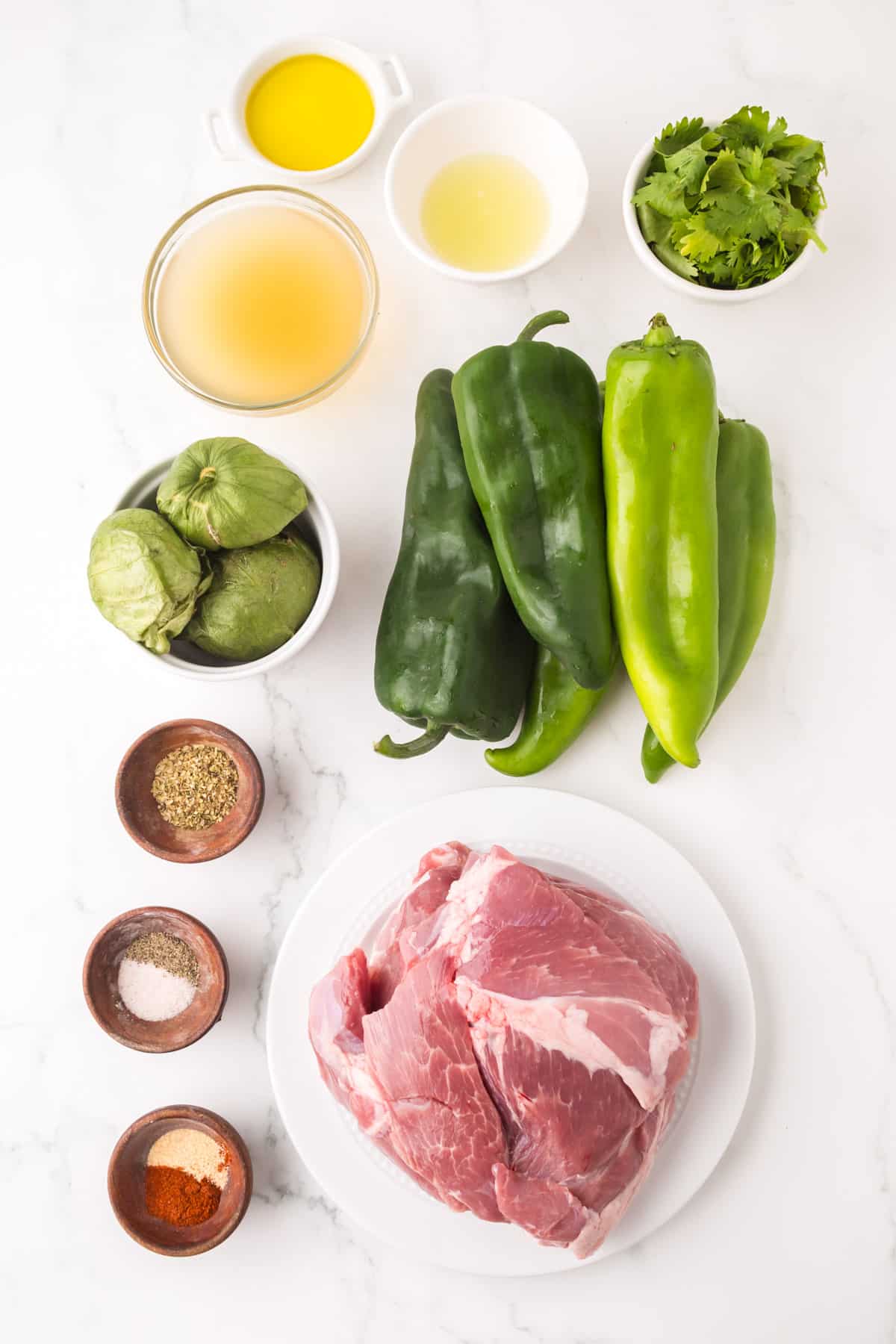 Overhead view of raw pork on a plate, green chilies, tomatillos, fresh cilantro, olive oil, broth, lime juice, and small bowls of spices arranged on a white marble surface.
