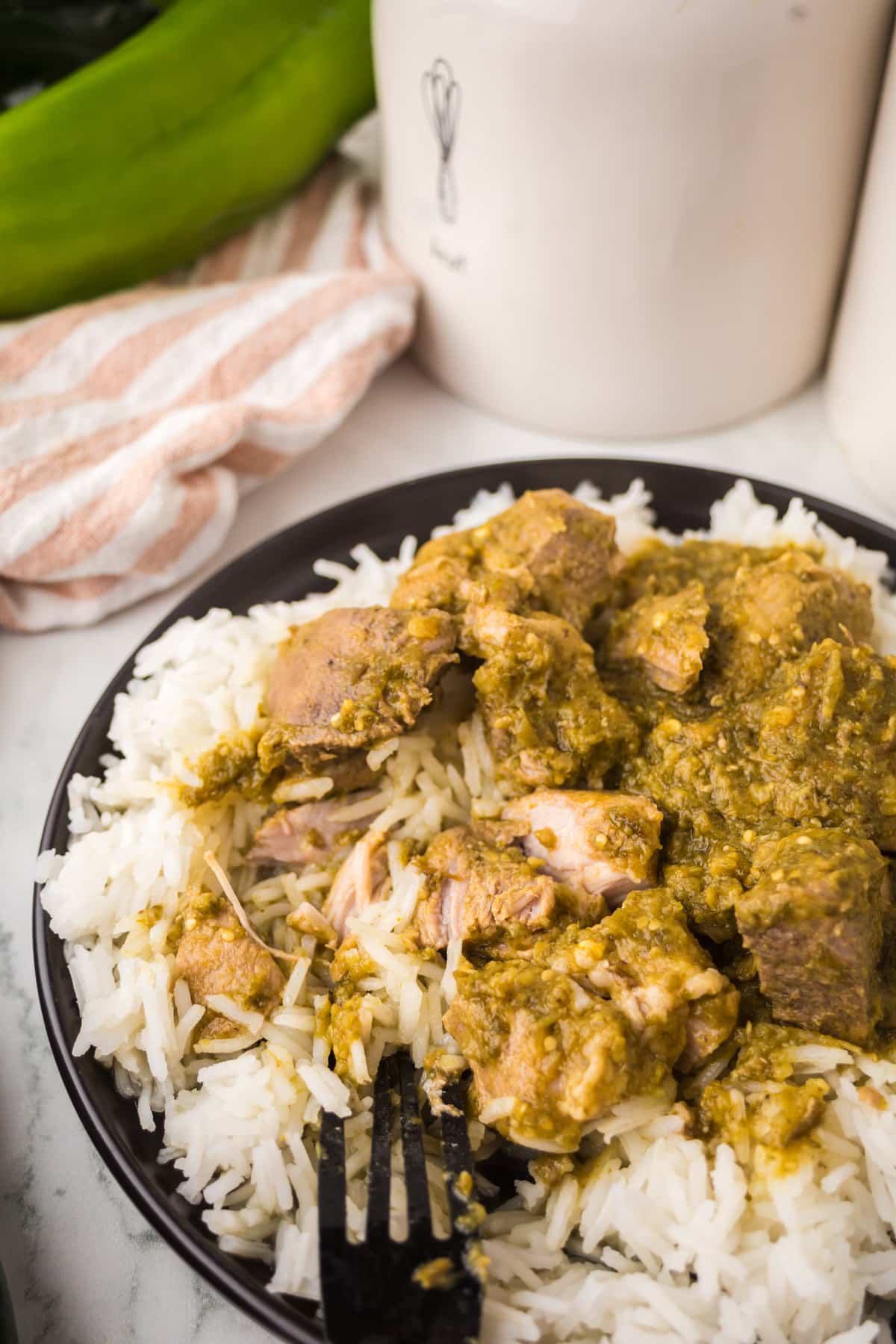 A black plate filled with white rice topped with chunks of meat covered in green sauce, with a black fork resting on the plate. A striped cloth and ceramic containers are in the background.
